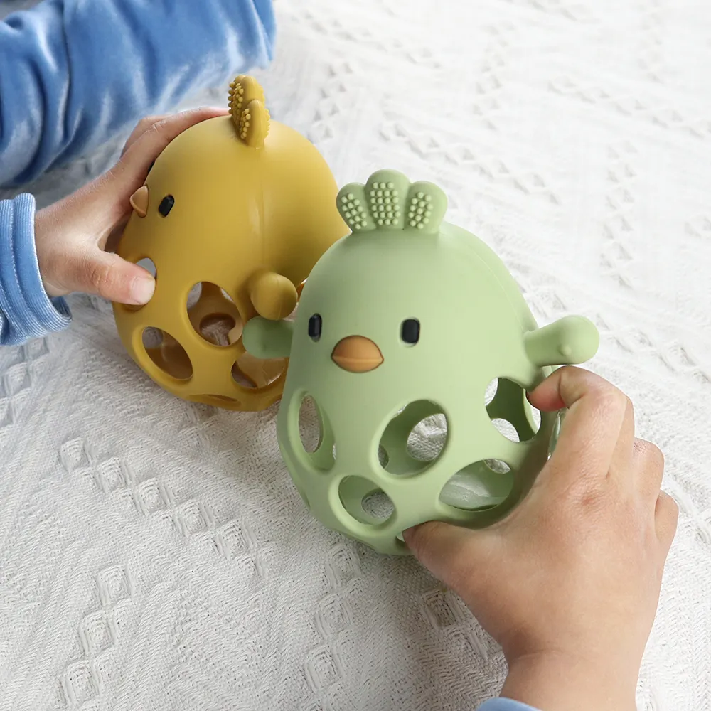 Baby holding yellow and sage green chick-shaped silicone teether rattles on white textured blanket.